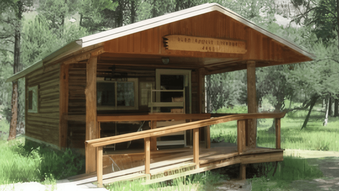 A small wooden cabin with a covered porch and wheelchair-accessible ramp sits surrounded by trees and grass in a sunlit forest clearing.