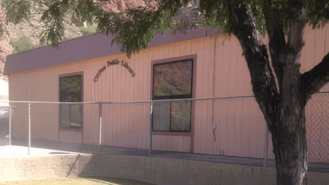 A light pink building with large windows and a chain-link fence in front. A tree with green leaves partially shades the building. The setting appears to be outdoors on a sunny day.