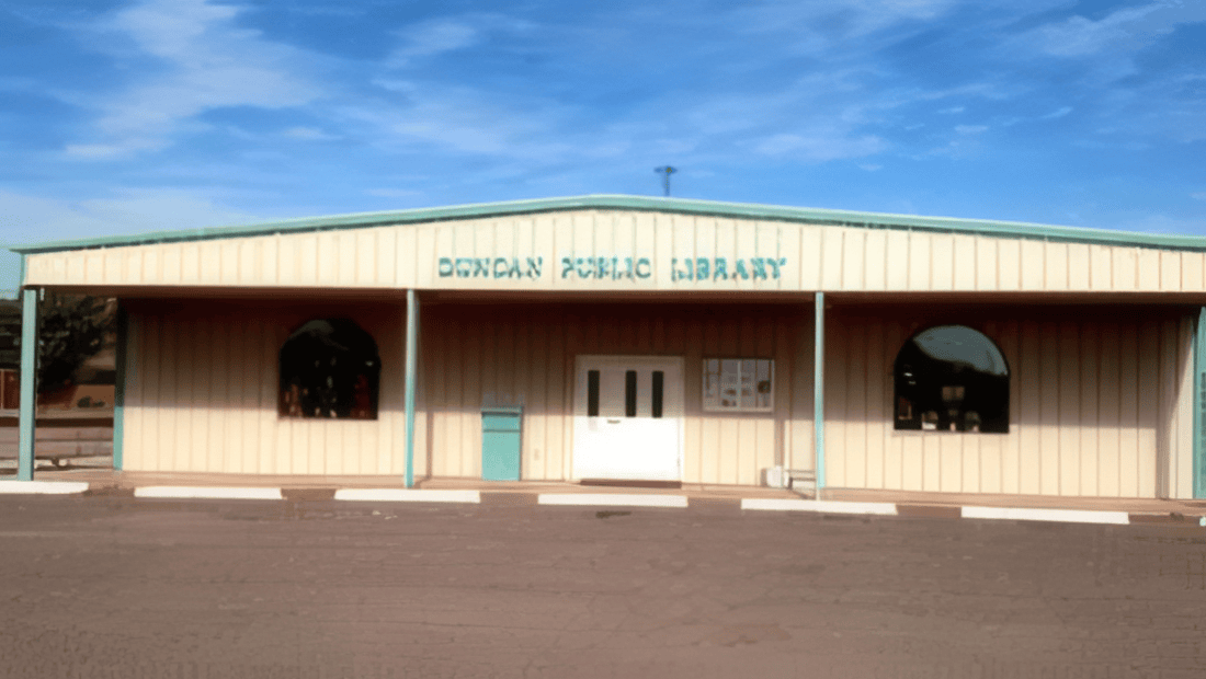 A beige, metal building labeled “Duncan Public Library” stands under a blue sky, with arched windows and white double doors at the front. White parking barriers are visible in front of the entrance.